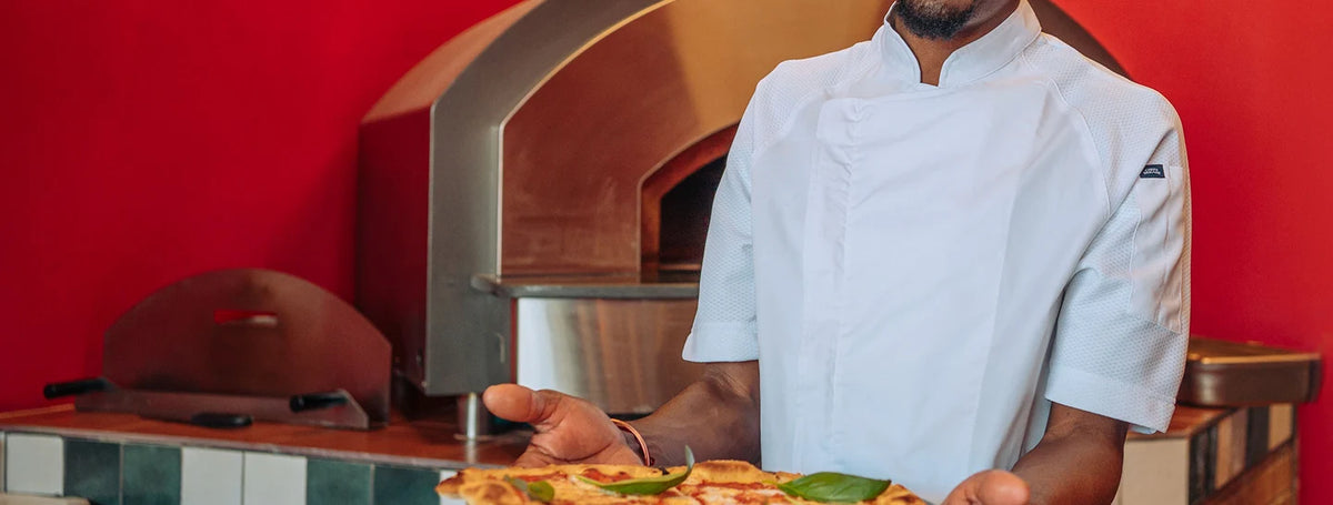 A chef in a white uniform holding a freshly baked pizza in front of a red wall and wood-fired pizza oven.