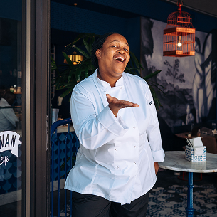 A smiling chef in a white double-breasted jacket stands in a restaurant setting, gesturing with their hand.